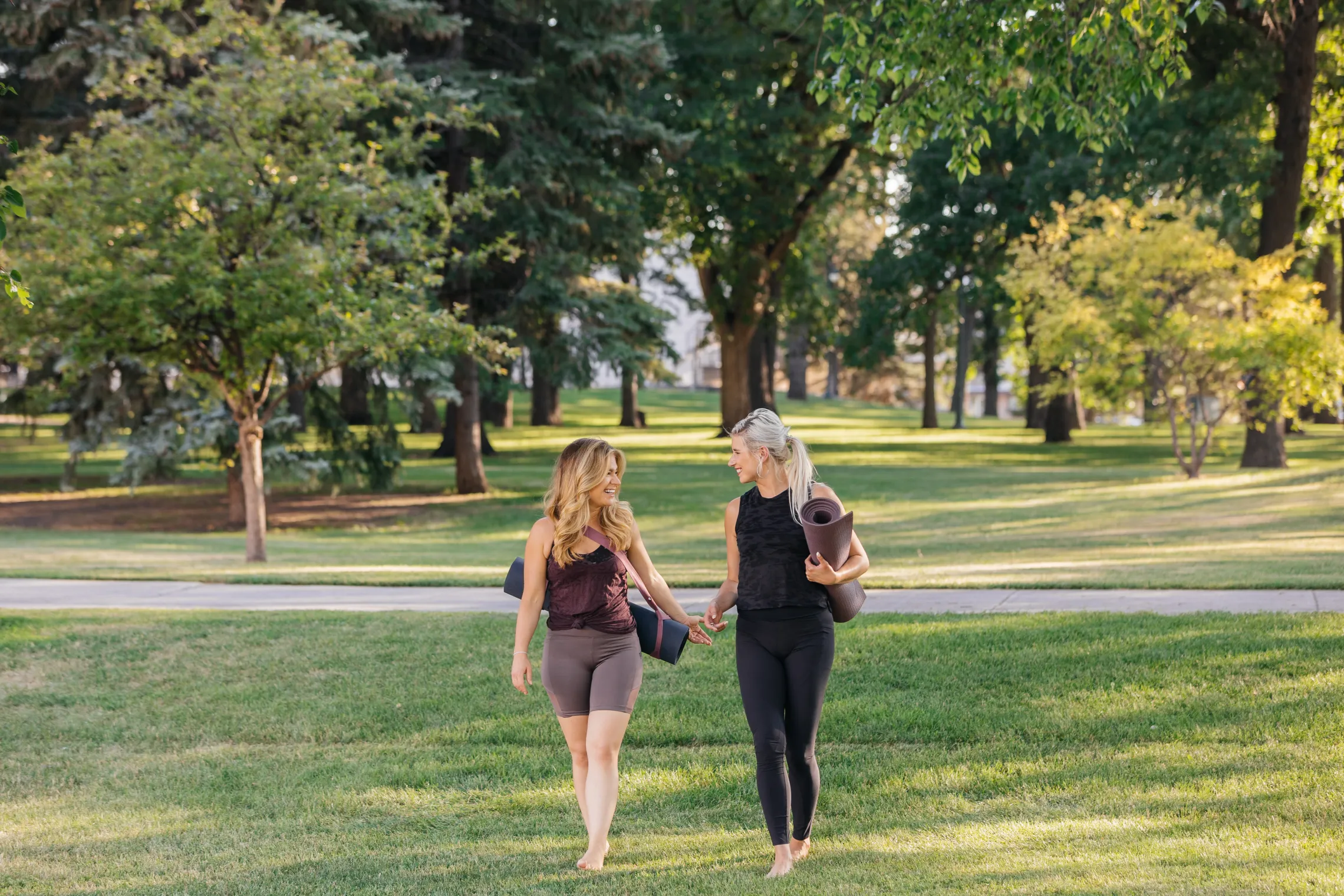 Two women chatting as they walk through a park with their yoga mats.