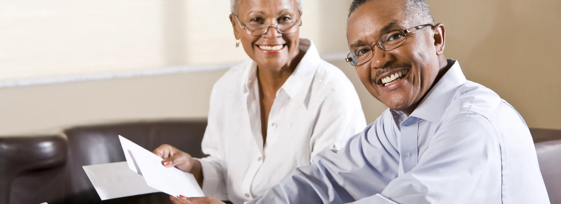 Two older adults sit together on a couch, smiling while reviewing paperwork, with one holding an envelope and documents.